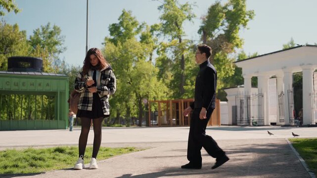 Woman stands outdoors holding phone while looking down as another person approaches. setting includes outdoor elements like buildings and nature man conversation