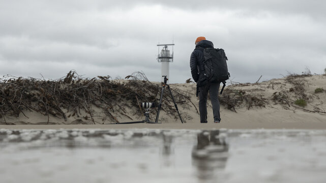 View of a photographer with a backpack and tripod standing on the sandy beach with a lighthouse in the distance under a cloudy sky in Debki, Pomeranian Voivodeship, Poland.
