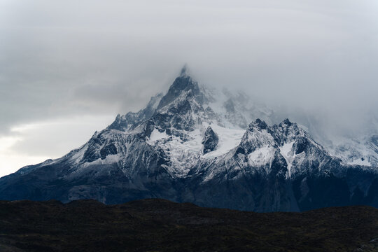 View of Cordillera del Paine mountain peak covered in snow and shrouded in low-hanging clouds above dark rocky terrain in Torres del Paine, Magallanes, Chile.