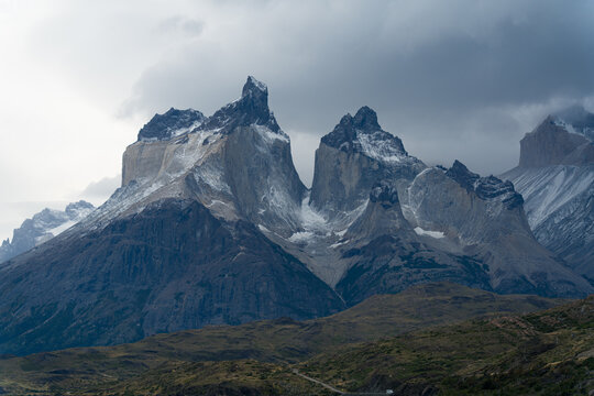 View of the jagged granite peaks of the Cordillera del Paine rising above the rugged landscape under a dramatic cloudy sky in Torres del Paine, Magallanes, Chile.