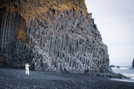 View of a woman in a white dress standing on the black sand of Reynisfjara Beach in front of towering basalt columns in Vik, Myrdalshreppur, Iceland.