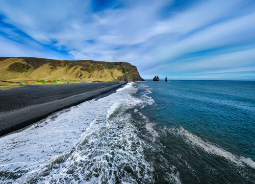 View of Reynisfjara Beach with its iconic black sand, crashing ocean waves, and dramatic sea stacks rising from the water under a bright blue sky in Vik, Myrdalshreppur, Iceland.