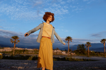 A woman in a striped top and mustard skirt balances gracefully outdoors, denim jacket fluttering, against a sunset sky with palm trees and open landscape. © SHOTPRIME STUDIO