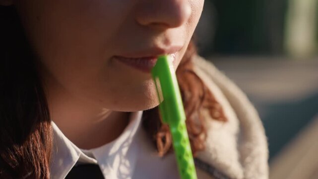 Close up view of lower face profile showing woman lips partially obscured by bright sunlight. Visible curly hair and white shirt with black tie suggest school or work setting. Green pen held