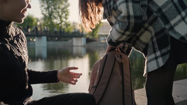 Two people interact near calm water during sunset. One person stands to hand over backpack while other sits receiving objects in intimate outdoor setting