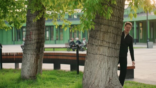 Man dressed in all black slowly appears between two large leafy tree trunks revealing public space with wooden benches on paved ground and green buildings in background. scene features soft