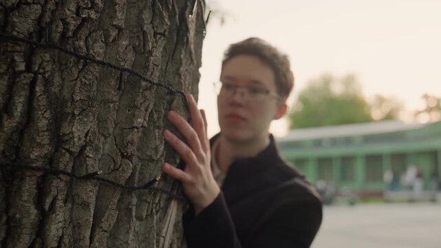 Close up focus on textured surface of large tree bark is shown. Hands gently touch or adjust string lights wrapped around trunk with soft outdoor lighting and blurred background elements