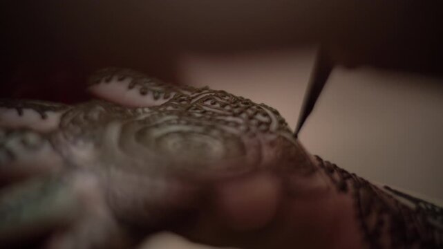 The tip of a henna cone draws fine lines over a detailed mehndi pattern on the back of a hand, with the focus shifting across the design, Macro
