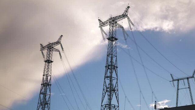 Low angle view of steel transmission towers carrying multiple power lines as clouds drift across a bright blue sky above an industrial landscape