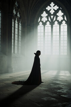 Vertical photo of a man in a long black and red cape standing in a gothic cathedral. Dramatic silhouette of a vampire in a misty stone hall with light rays shining through arched windows