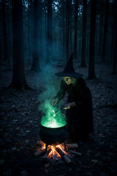 Vertical illustration of an old witch brewing a magic potion in a glowing green cauldron. Spooky woman casting a spell in a dark forest at night. Halloween witchcraft concept