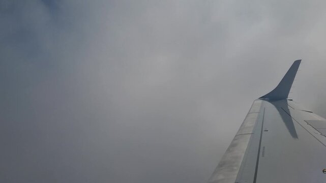 Airplane window view of the wing passing through layers of white and gray clouds during descent toward Serbia, capturing changing weather conditions and a scenic aviation journey