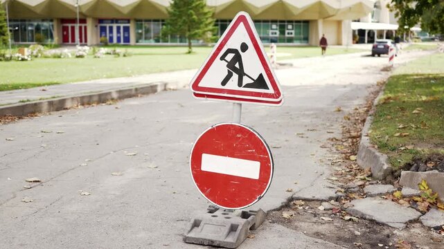 Road work warning signs displayed on a city street, featuring a no entry symbol and surrounded by grass and contemporary architecture in the background