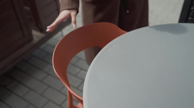 Woman approaches outdoor setting with tables and chairs. Woman pulls out chair sits down at table then places coffee cup on it before picking up her phone