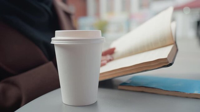 Shows person holding hands near books and cups in soft focus. white disposable coffee cup sits on table next to open book with hand of someone wearing pink nail polish with star designs