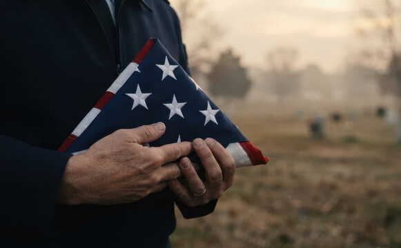 A male military veteran in dark uniform holding a folded American flag at a cemetery for a solemn funeral