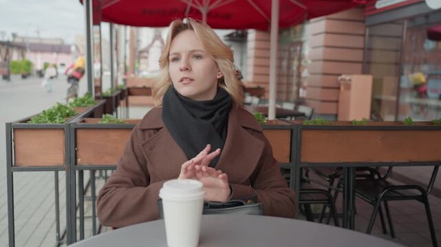 Lady is seated outdoors in public area possibly near street or cafe. Woman appears to be waiting for something while fidgeting with her hands and looking around environment