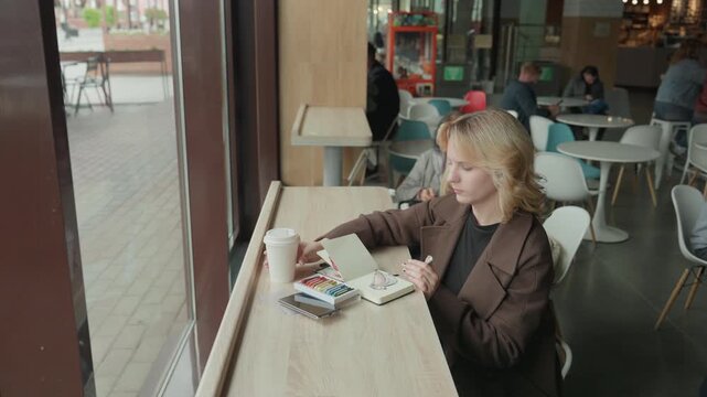 Woman sketches with art supplies at cafe table. Woman uses pencils on paper while surrounded by other patrons indoors. scene features coffee cups and artistic equipment in natural lighting