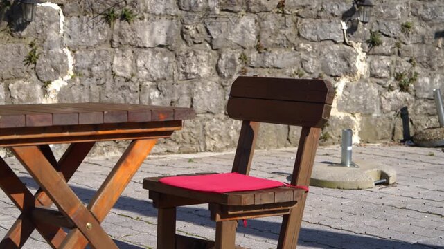 A folding wooden table and a chair with a red cushion stand empty on a cobblestone patio beside an old rough stone wall, as the camera drifts slowly past an outdoor cafe seating area.