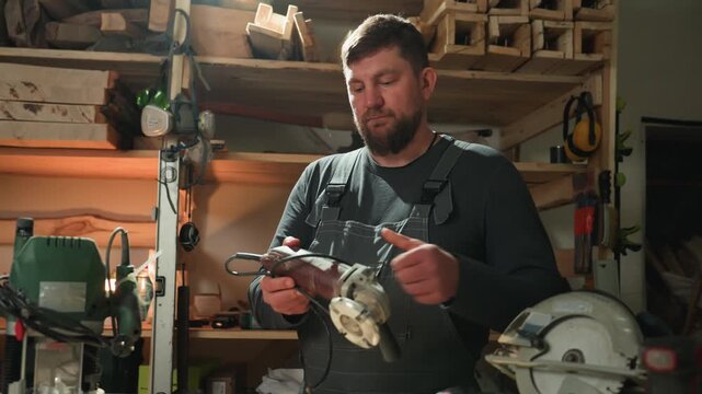 Carpenter works at his bench using various hand tools including saws. workshop is filled with wooden planks green router machine on table and other woodworking equipment in shelves