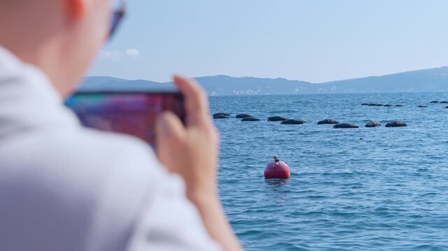 A man in a light shirt holds a smartphone and films floating mussel farm lines and a red buoy on calm blue water, with distant hills on the horizon, a concept of coastal aquaculture tourism.