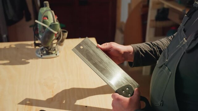 Craftsman examines or prepares to use large handsaw with workshop elements like power tools visible in background. scene focuses on woodworking preparation and craftsmanship