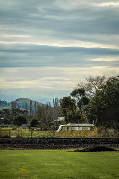 A weathered yellow bus rests among vegetation by railway line in a small rural town near rolling hills, against vast layered sky, Terang, Victoria, Australia