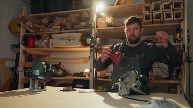 Man works at wooden workbench handling power woodworking tools like sander and saw. background shows shelves filled with wood planks and workshop supplies