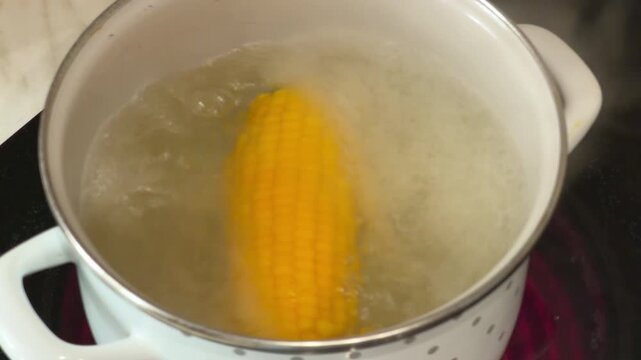 A yellow sweet corn cob cooks in rolling hot water inside a white enamel saucepan with polka dots on an electric hob, a simple home recipe for a healthy vegetable meal.