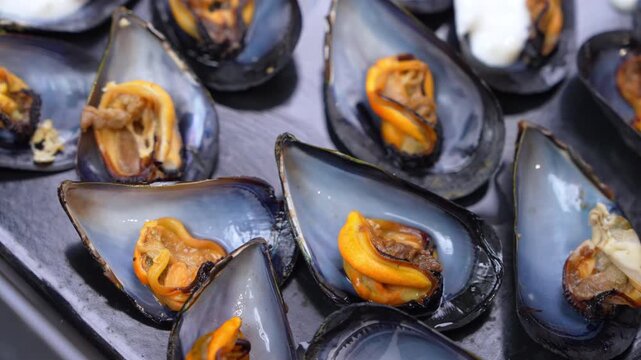 Steamed mussels with orange meat rest in open blue black shells on a slate platter, some topped with creamy white sauce, a tasty seafood dish for restaurant menus and gourmet cooking.