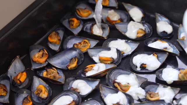 A stream of creamy white sauce falls onto rows of mussels resting in open shells on a dark metal tray, a catering kitchen step for banquet seafood service.