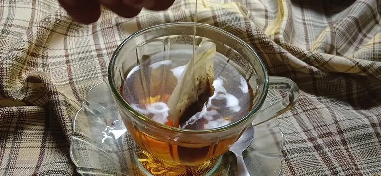 hand dips the tea bag in a transparent glass cup with warm water in it which turns brown slowly