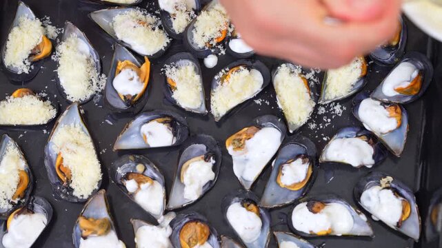 A hand uses a spoon to sprinkle grated cheese onto rows of mussels topped with creamy white sauce on a dark baking tray, a home baking step before the oven.