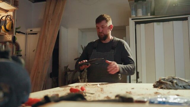 Man wearing dark clothing stands at workbench covered with wood shavings. Man interacts with various carpentry tools like screwdrivers and clamps examining them closely while surrounded