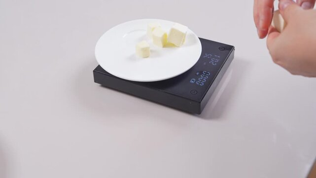 A hand drops small cubes of butter one by one onto a white plate resting on a black digital scale on a light countertop, as the weight reading climbs, a concept of precise baking measurement.