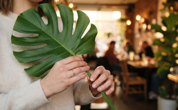 Young woman holding a large tropical monstera leaf in a cozy cafe interior for lifestyle content