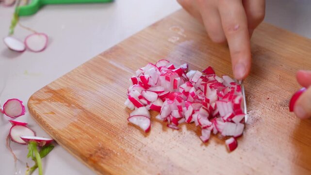 Hands with a knife gather finely diced red radish into a heap on a bamboo board, trimmed stalks resting on the counter, a concept of plant based batch cooking at home.