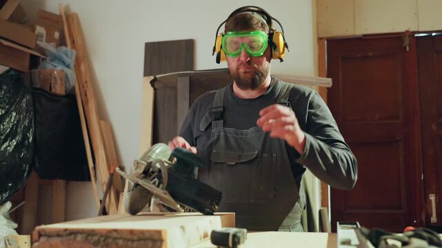Man operates circular saw on wooden plank inside cluttered workspace with various materials and tools visible. Man wears safety goggles and ear protection while handling power equipment