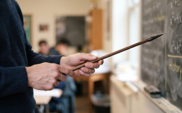 Male teacher pointing at mathematical formulas on blackboard with pointer stick in school classroom