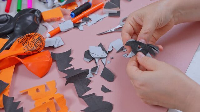 Close view of hands trimming a gray and black paper bat with narrow scissors over a pink surface strewn with offcuts, balloons and markers nearby, a seasonal home activity for kids.