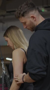 Male personal trainer gently guides a female client's upper arm during a cable rope triceps pushdown in vertical slow motion.