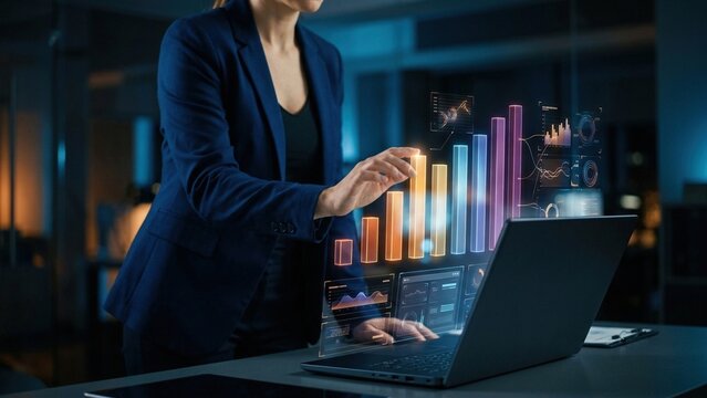 Businesswoman interacting with futuristic holographic financial data charts and graphs on a modern office desk with a laptop analyzing performance.
