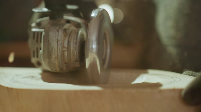 Close up shows electric hand held sanding machine precisely carving circular pattern into smooth surface of light colored wood with fine dust particles visible as it cuts. background remains