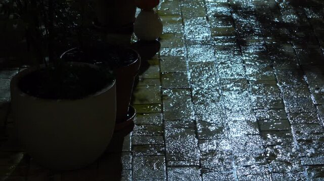 Raindrops splash and shimmer on a wet paved stone path next to large ceramic planters in a dark outdoor courtyard, a moody rainy evening scene with reflective light on the stones.