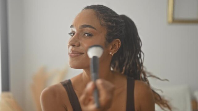Woman applying makeup brush to cheek in studio, smiling and holding brush to face while looking at camera; confidence.