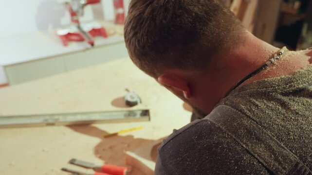 Close up shows back of man head as man works on detailed wood carving with chisel and hammer in workshop covered in sawdust. scene includes woodworking tools like level ruler pencil