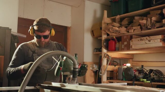 Man uses handheld router to shape wooden material on workbench. Man wears protective gear including ear muffs gloves glasses in workshop filled with tools and lumber