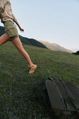 Naklejka premium Young woman in khaki shorts leaps across a sunlit meadow with a backpack on the ground, mountains on the horizon, conveying carefree outdoor adventure and movement.