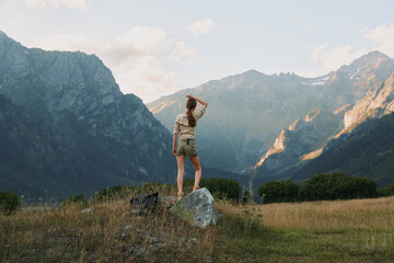 Naklejka premium Young woman stands on a rock in a sunlit mountain meadow, gazing toward distant peaks with a sense of freedom and calm, capturing a natural outdoor moment.