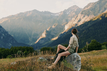 Naklejka premium One woman sits on a rock in a mountain meadow, gazing toward distant peaks, conveying calm, freedom and solitude in a pristine outdoor scene.